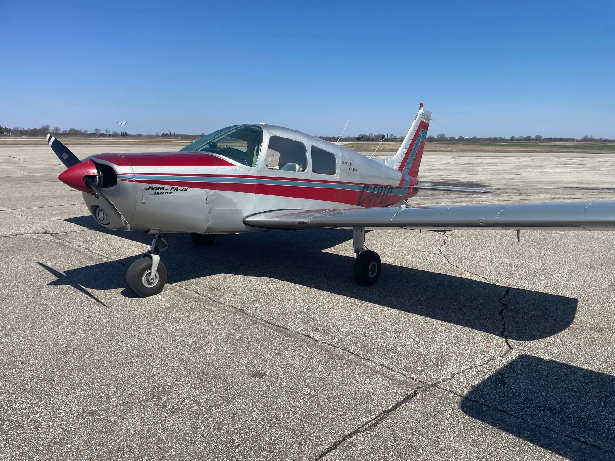 A silver and red Piper PA-28 single-engine aircraft, registration C-GOMF, parked on an airfield tarmac under a clear sky.