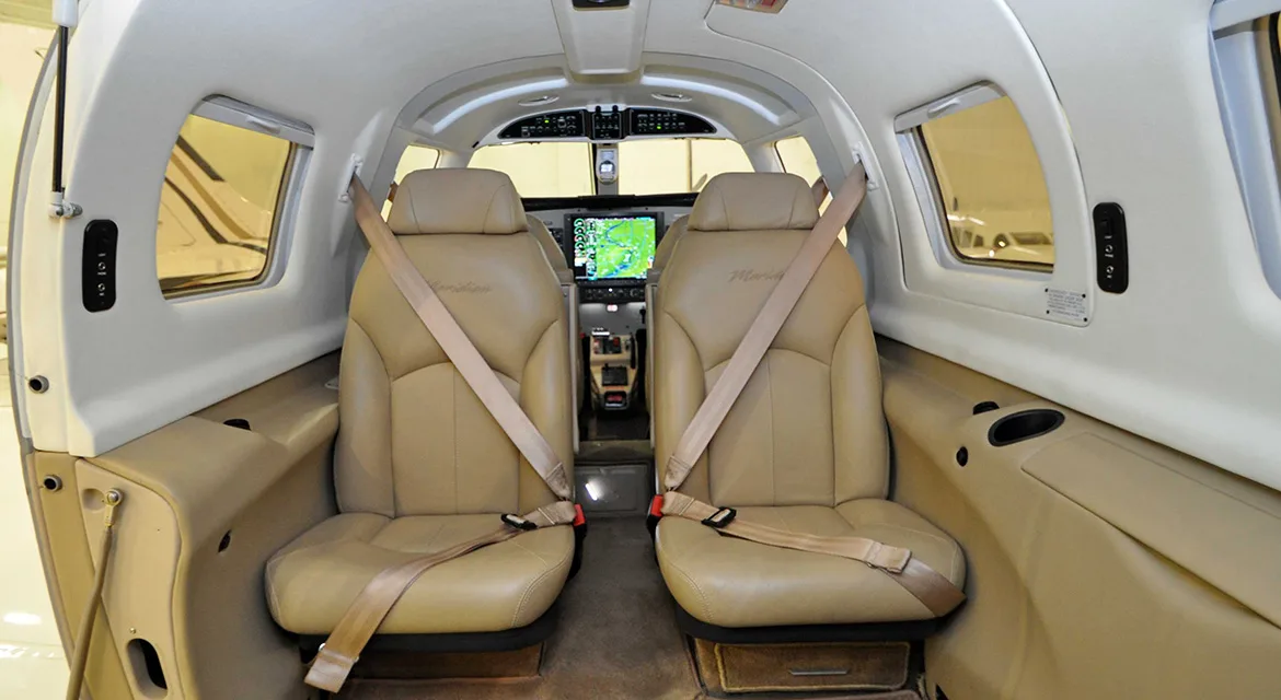 The tan leather interior of an aircraft cabin, showing two front passenger seats with seatbelts and a glimpse of the cockpit.