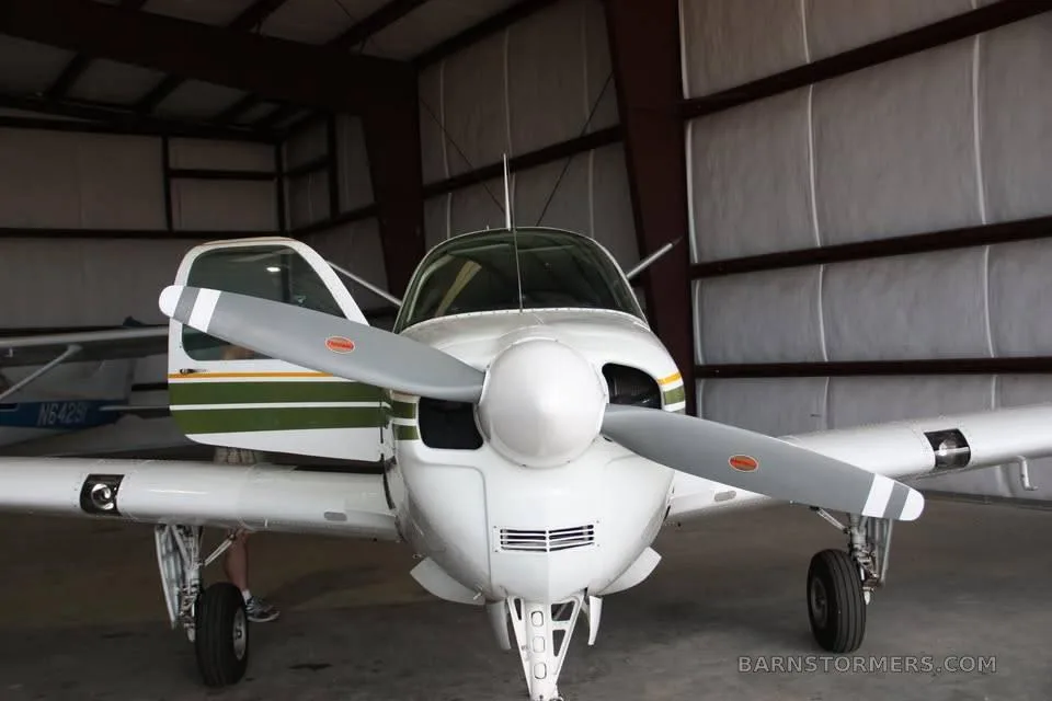 A front view of a white, green, and yellow single-engine propeller aircraft with its door open, parked inside a hangar.