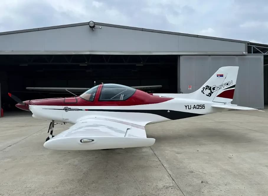 A white and red low-wing Griffon aircraft with registration YU-A056 parked on a tarmac in front of a hangar.