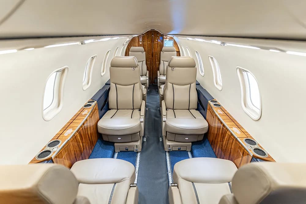 The aircraft cabin interior with tan leather seats, wood trim, and blue carpet.