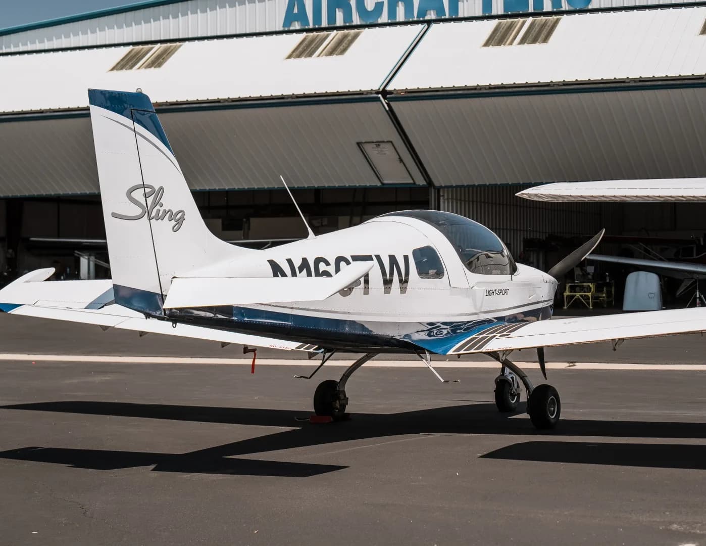 A white and blue Sling Light-Sport aircraft, N166TW, parked on an asphalt apron with a hangar in the background.