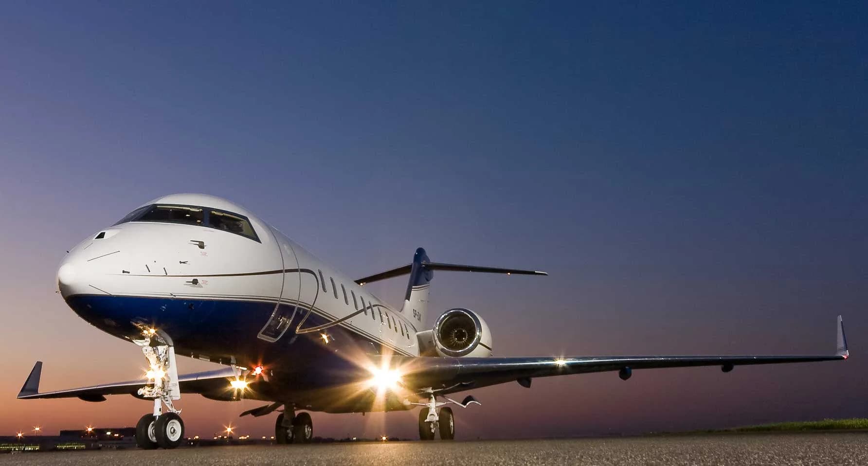 A white and blue private jet with its landing lights on, parked on an airfield at dusk.