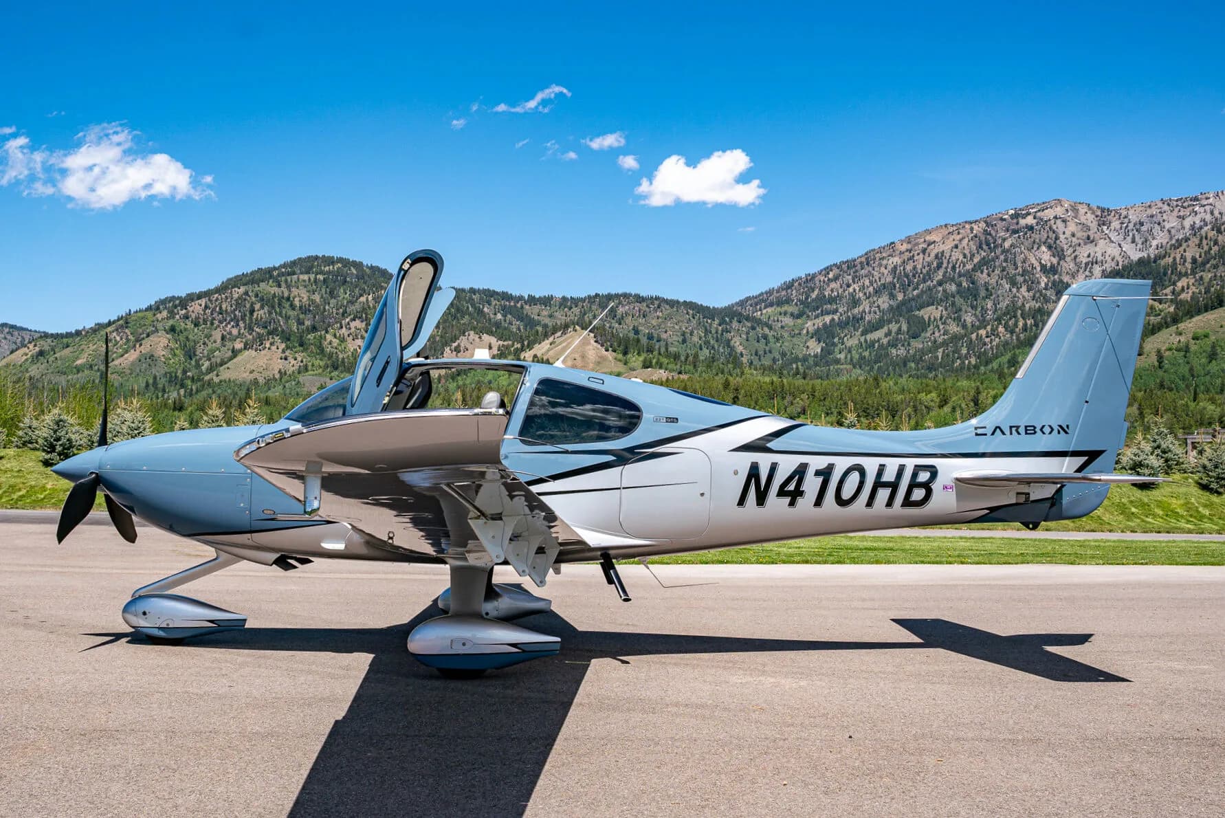 A light blue and silver Cirrus SR22T aircraft (N410HB) with its doors open, parked on an airfield with mountains in the background.