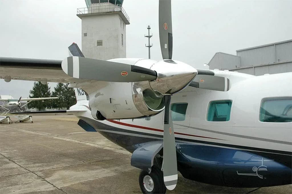 A close-up of the right engine and fuselage of a white and blue twin-engine turboprop aircraft, with a control tower in the background.