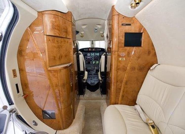 The luxurious interior of a private jet cabin features light leather seats, polished wood cabinetry, and a view into the cockpit.