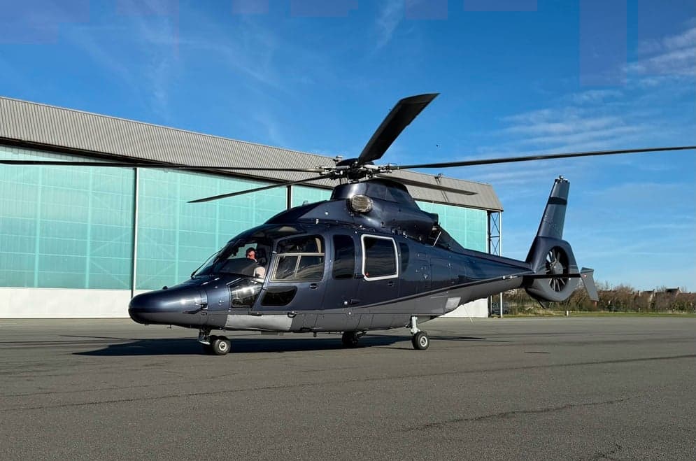 A dark blue helicopter is parked on an airfield next to a large hangar under a clear blue sky.