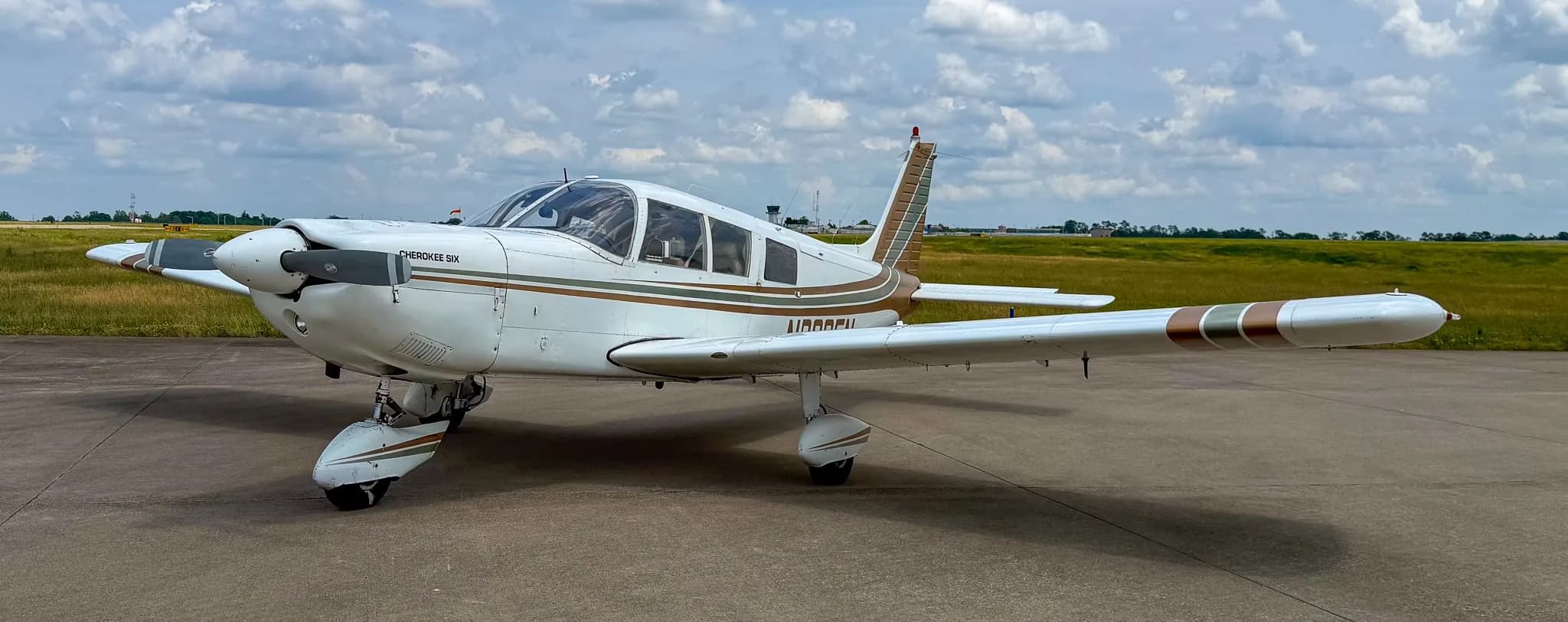 A front-right view of a white Piper Cherokee Six aircraft with brown and green stripes on an airfield under a cloudy sky.