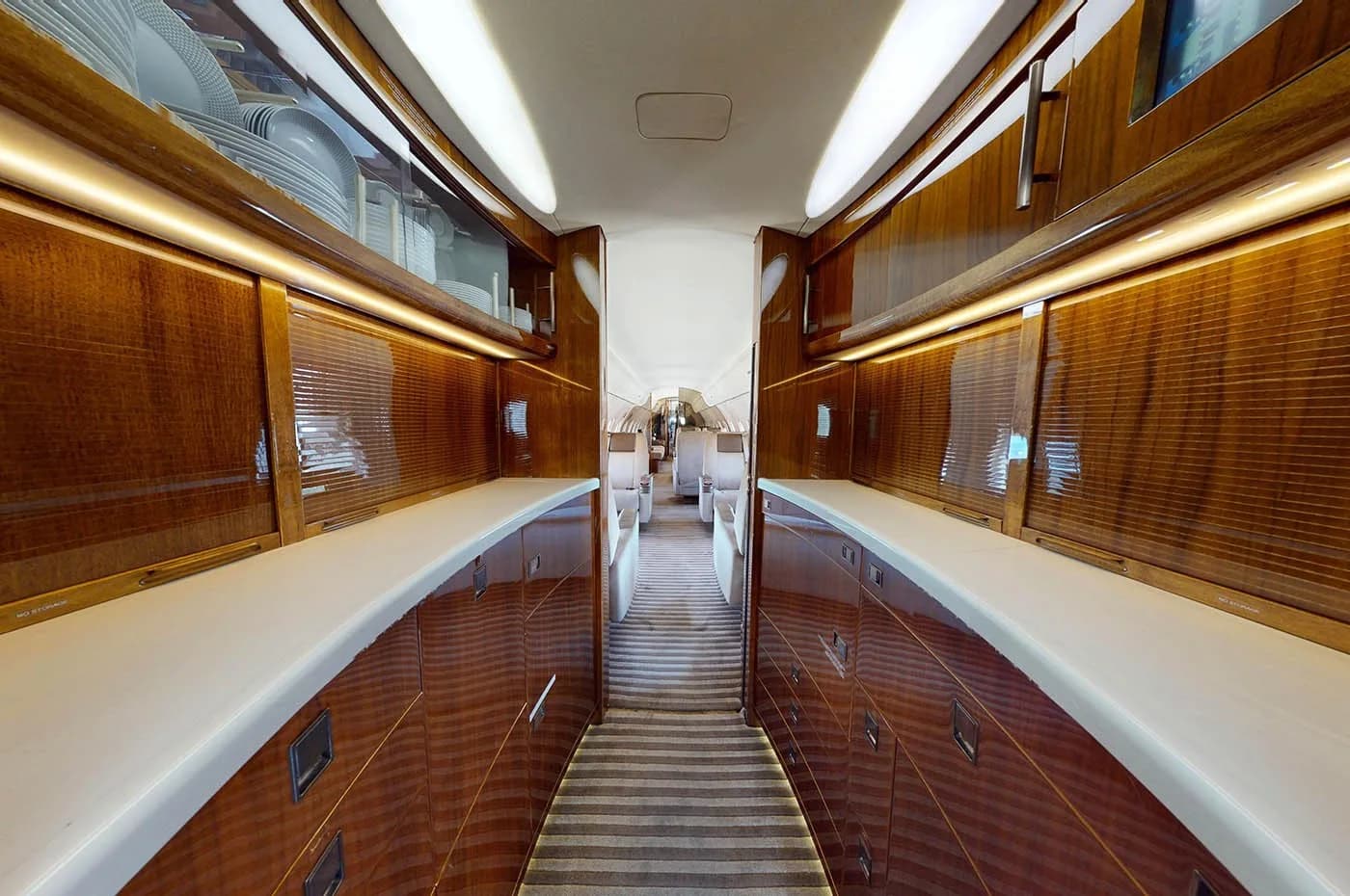 The galley area of a private jet, featuring polished wooden cabinetry, white countertops, and overhead storage with dishes.