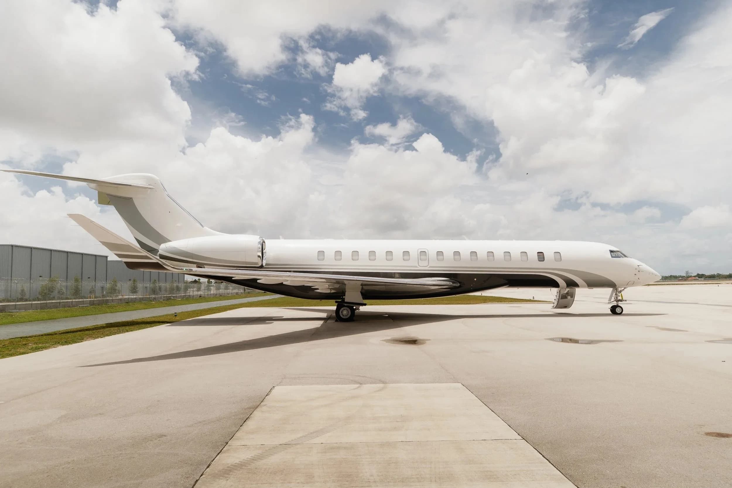 A white and grey Bombardier Global 7500 business jet is parked on an airfield under a cloudy sky, viewed from the side.