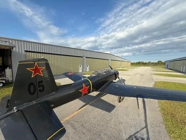 An aerial-like view from the rear of a dark gray Yak-52 aircraft with red stars and yellow accents parked on an asphalt ramp.