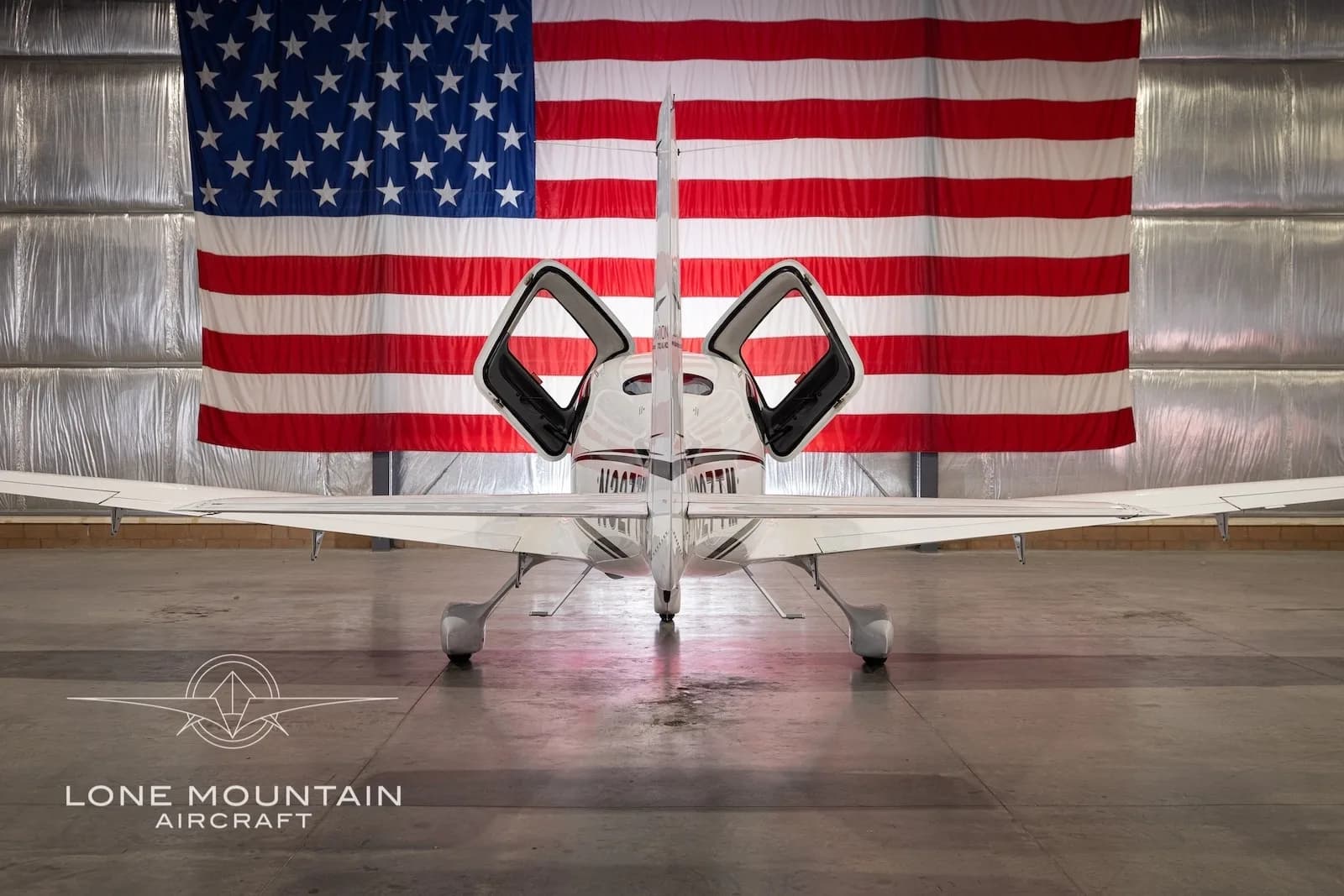 Rear view of a white Cirrus SR22 aircraft with registration N327TM, parked in a hangar with an American flag backdrop, with its gull-wing doors open.