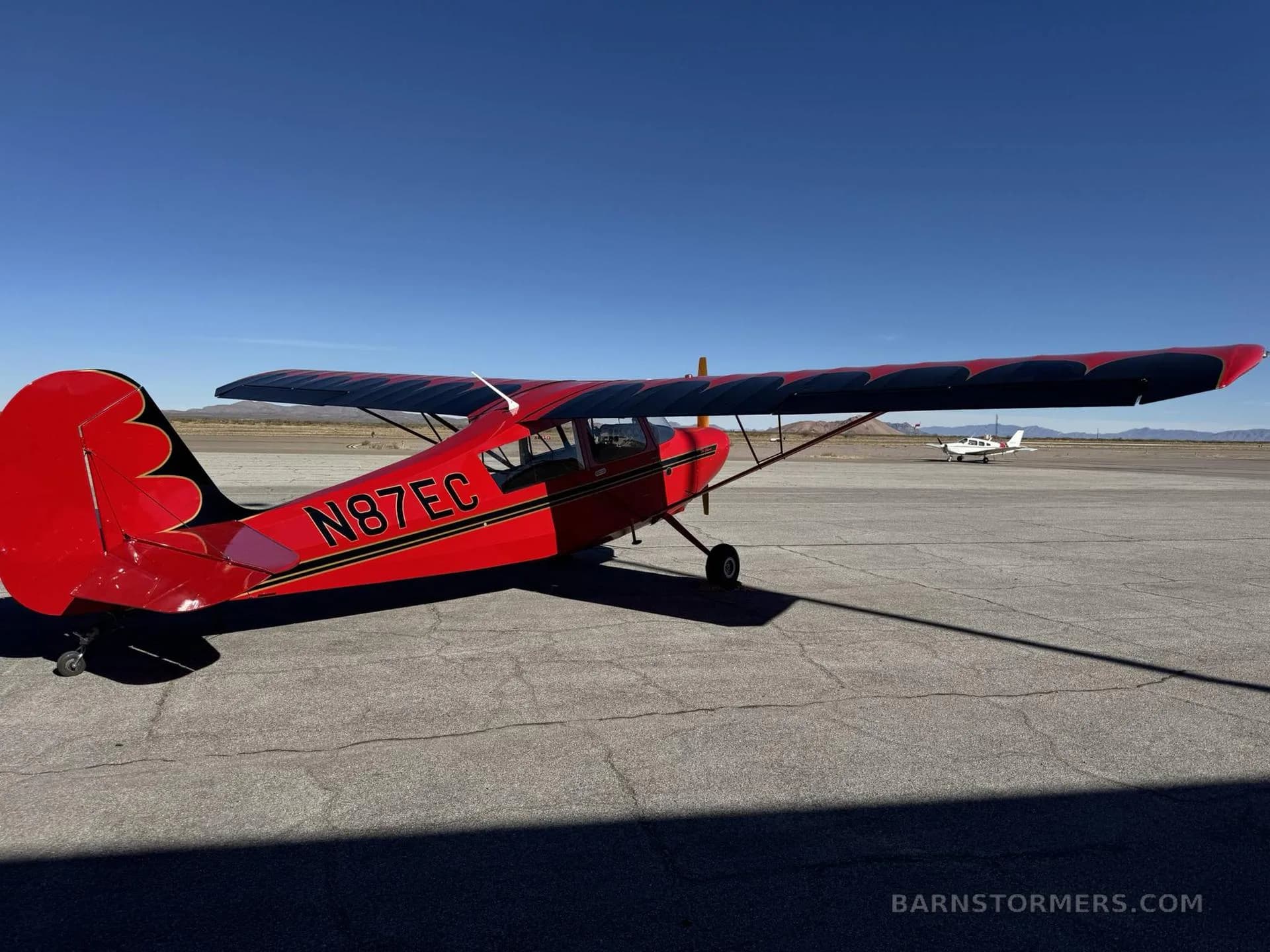 A red high-wing monoplane with registration N87EC is parked on an airfield, viewed from the rear-right, with another small aircraft in the distance.