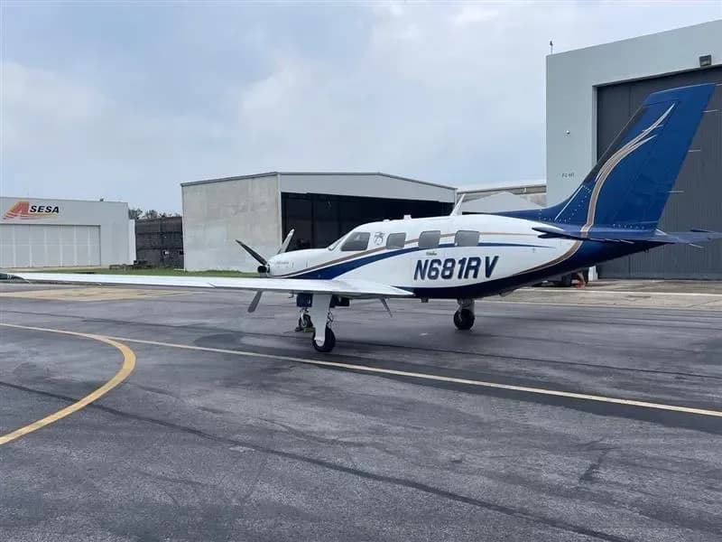 A white and blue twin-engine turboprop aircraft, N681RV, is parked on an airfield with hangars in the background under a cloudy sky.