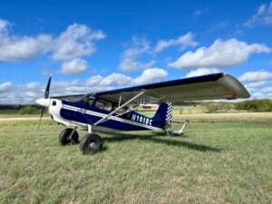 A blue and white ACA aircraft, registration N101BC, parked on a grassy field under a blue sky with scattered clouds.