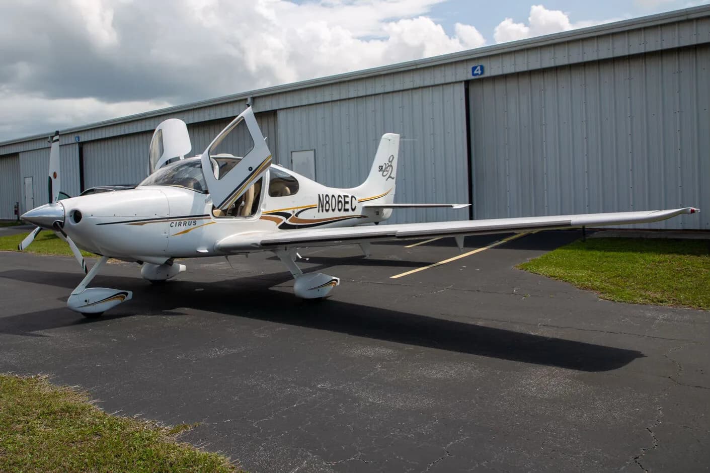 A white Cirrus SR22 aircraft, registration N806EC, with both gull-wing doors open, parked on an asphalt tarmac in front of a hangar.