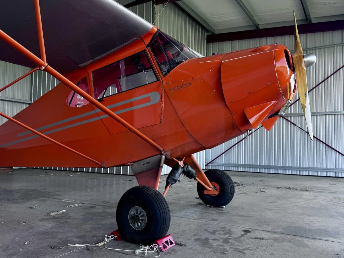 A front-right view of an orange vintage aircraft with a wooden propeller inside a hangar.