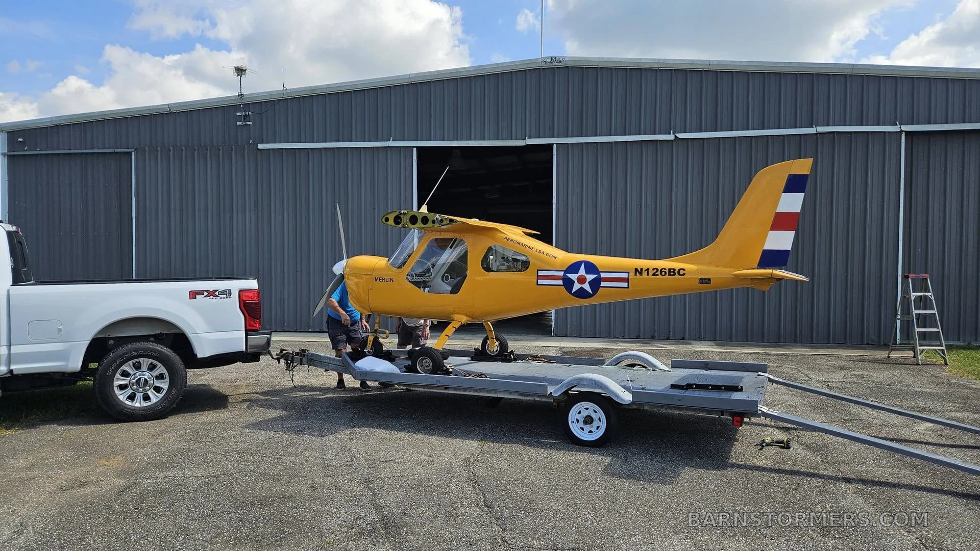 A yellow Merlin ultralight aircraft with a US star insignia is being loaded onto a trailer next to a white pickup truck and a gray hangar.