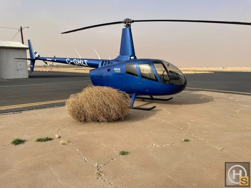 A blue Robinson R66 Turbine helicopter, registration C-GHLT, is parked on a concrete pad next to a tumbleweed.