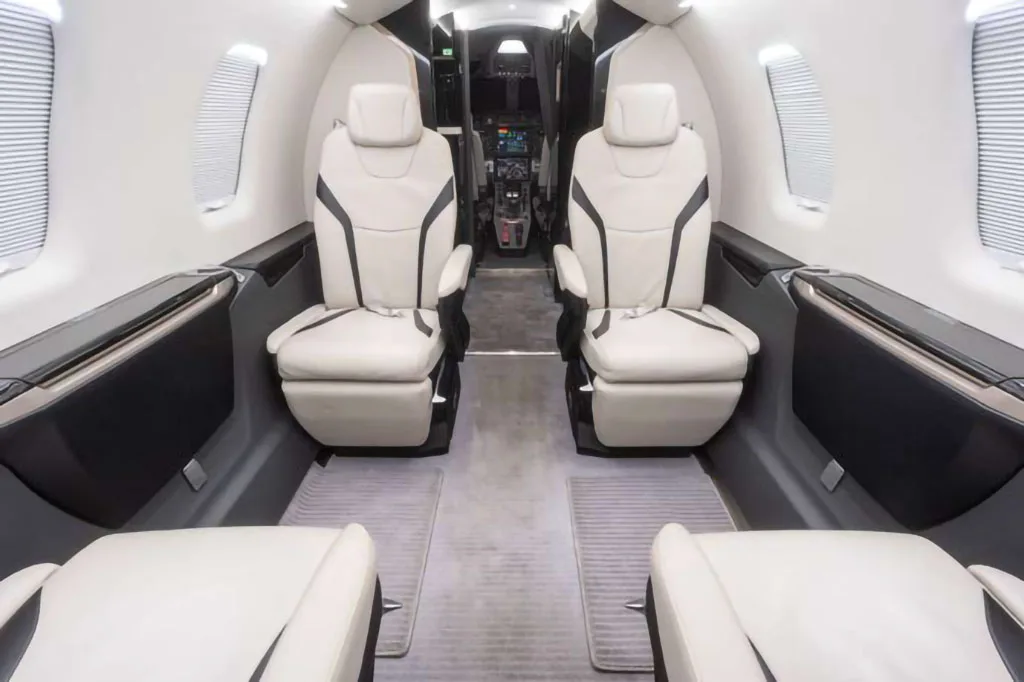 The luxurious interior cabin of a private jet features white leather seats with black accents, looking towards the cockpit.