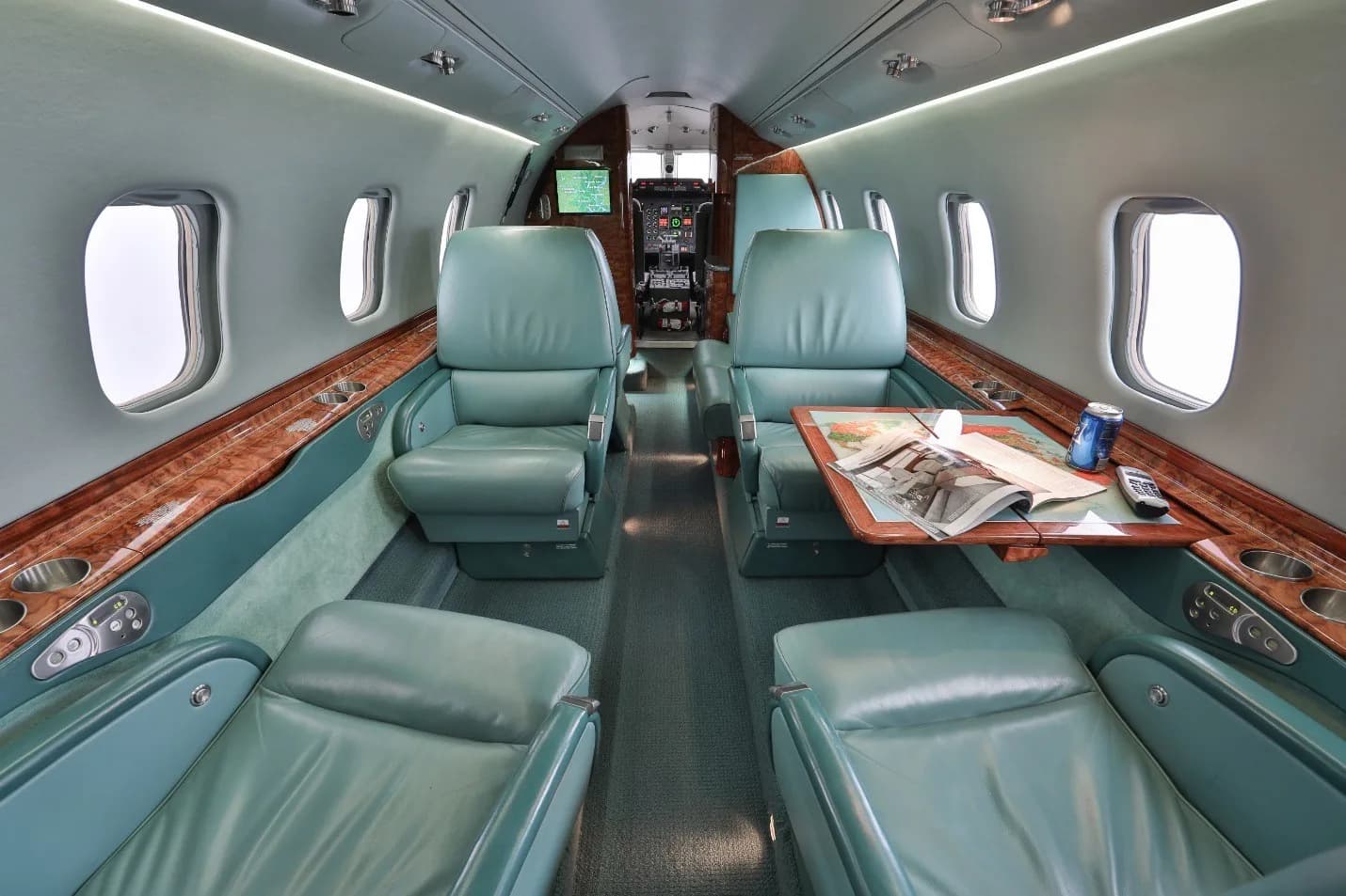 The luxurious interior cabin of a private jet with teal leather seats, wood paneling, and a table with a magazine.