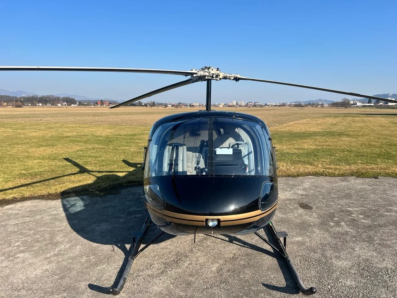 A front view of the black and gold Enstrom 480B helicopter (S5-HUA) on an airfield under a clear blue sky.