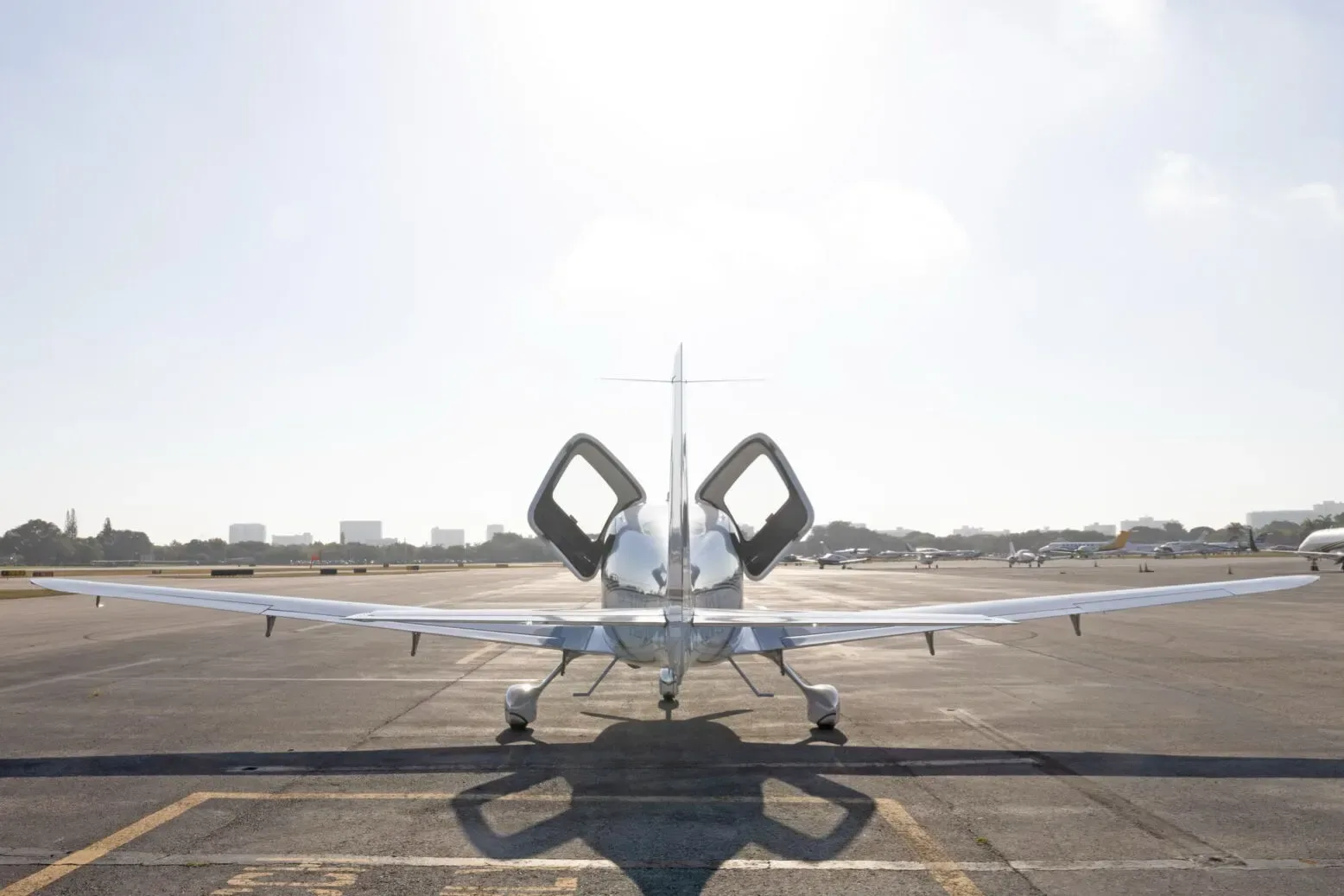 A white Cirrus SR22 aircraft is viewed from the rear with its doors open, parked on a sunny airport tarmac.