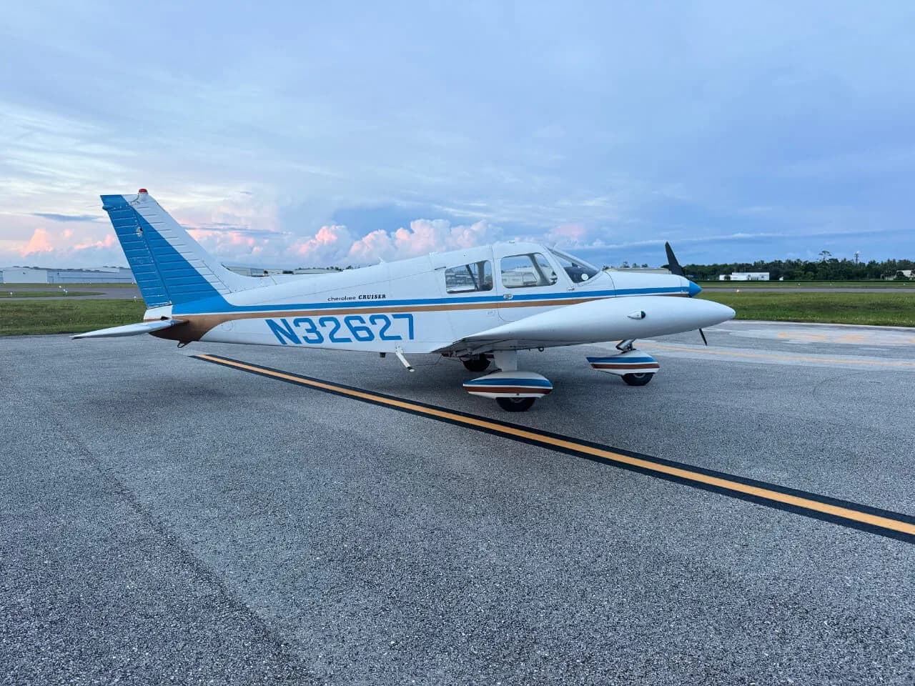 A white Piper Cherokee Cruiser with blue and brown stripes is parked on an airfield under a cloudy sky.