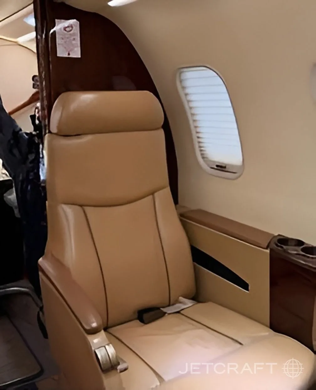 A single tan leather seat next to a window with closed blinds in the aircraft cabin.