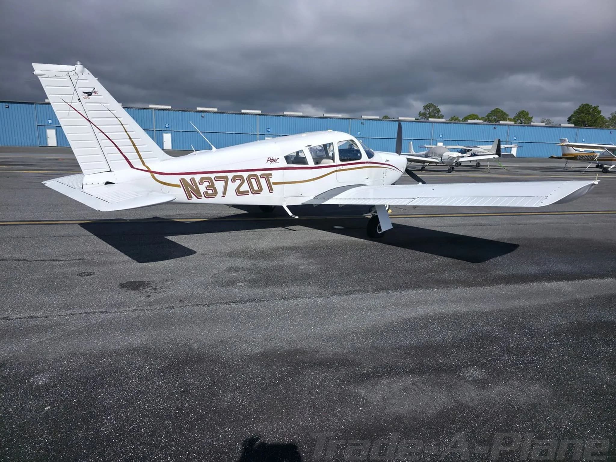 A side view of the white Piper Arrow N3720T parked on an asphalt apron with dark clouds overhead and hangars in the background.