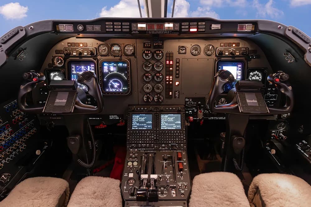 Detailed view of a private jet's cockpit with multiple screens, dials, and controls, and a blue sky visible through the windshield.