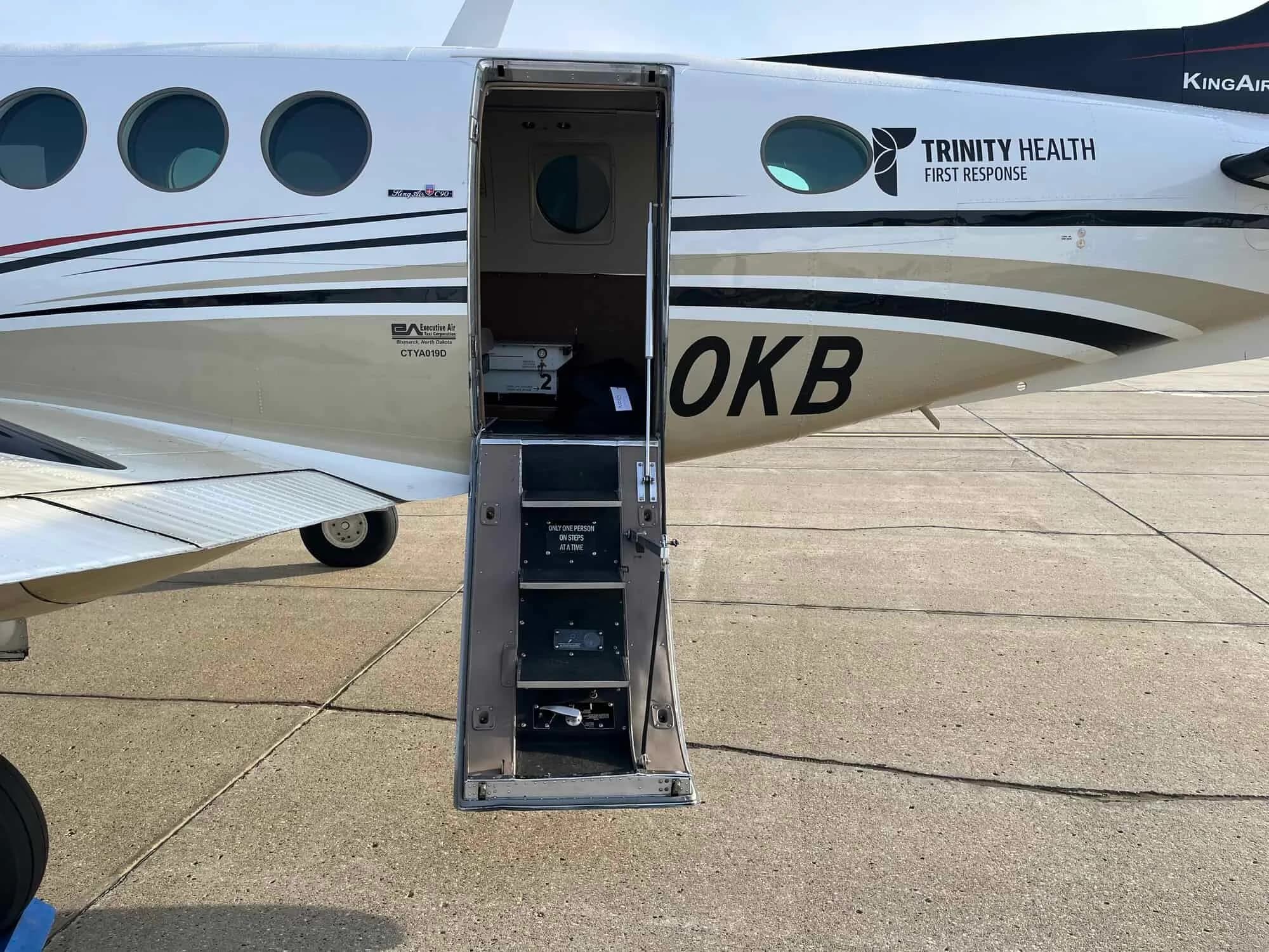 The open airstair door of a white and gold aircraft with "TRINITY HEALTH FIRST RESPONSE" text on the fuselage.