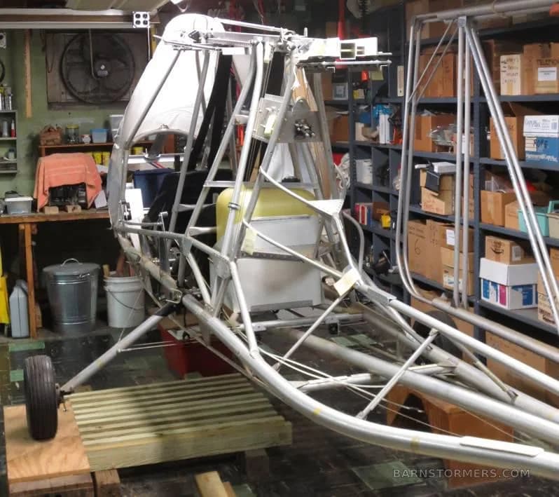 An overhead view of a partially assembled ultralight aircraft frame with a yellow fuel tank and black seats in a workshop.