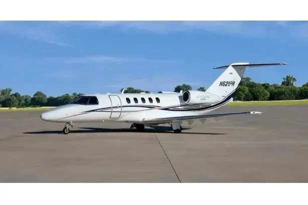 A white Cessna Citation CJ4 business jet with black and gold stripes, tail number N621PR, parked on an airfield under a blue sky.