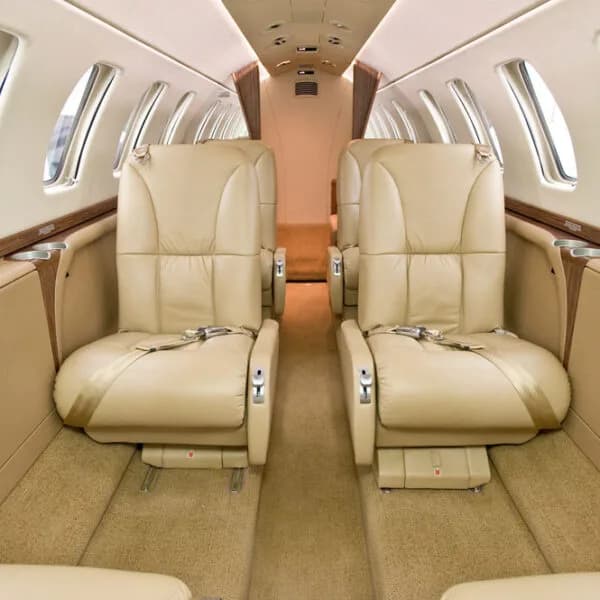 The interior cabin of a private jet, showing two rows of beige leather seats facing each other.