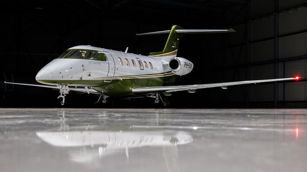 A white and green business jet, registration PH-DVK, parked in a dimly lit hangar.
