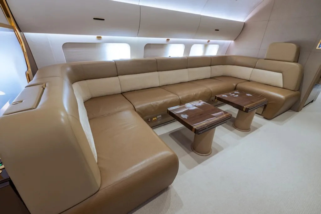 A comfortable L-shaped sofa in beige and brown leather with two small wooden tables in an aircraft's private cabin.