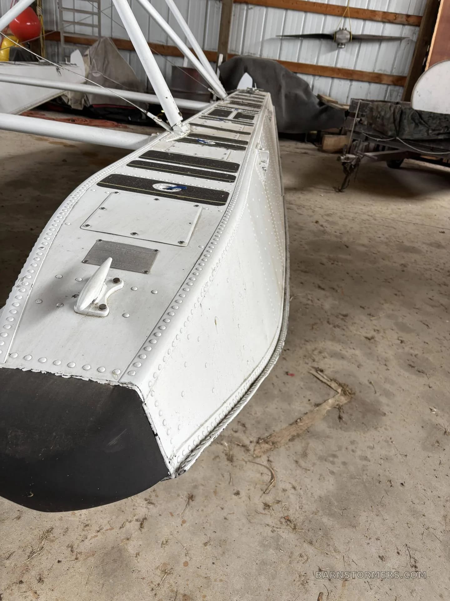 A close-up view of the front section of a white aircraft float with black anti-slip pads, showing rivets and a mooring cleat, inside a hangar.