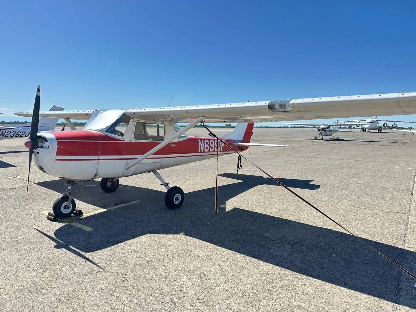 A red and white Cessna 150, tail number N6991F, parked on an asphalt apron with other aircraft in the background.