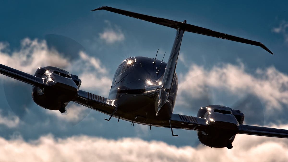 A dark-colored Diamond DA62 aircraft in flight against a cloudy sky.