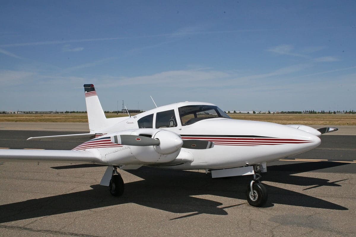 A white Piper Seneca twin-engine aircraft with red and blue stripes is parked on an airfield under a clear blue sky.