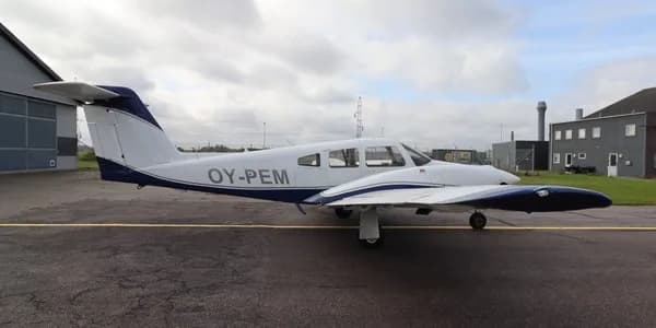 A white single-engine propeller aircraft with blue trim, registration OY-PEM, parked on an asphalt tarmac next to a hangar under a cloudy sky.