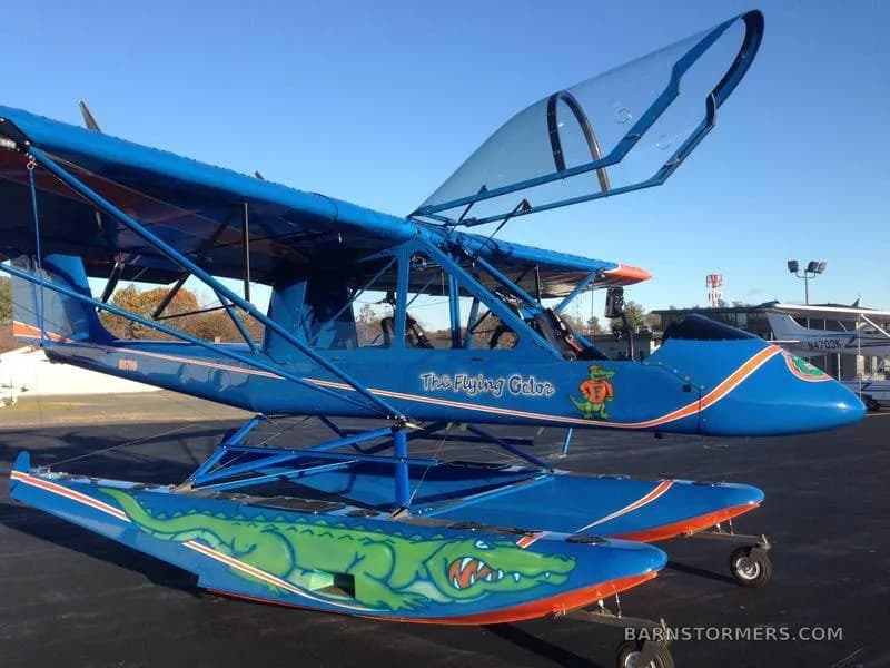 A blue floatplane with "The Flying Gator" painted on its side and a large green alligator graphic on its floats, parked on an tarmac.