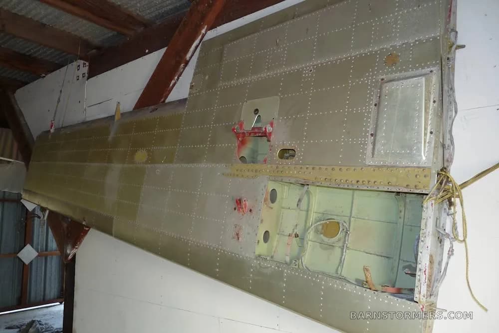 A close-up view of a riveted metal aircraft wing or tail section, showing internal structure, likely in a hangar.