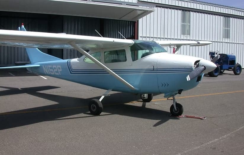 A light blue and white Cessna 152 is parked on the tarmac in front of a hangar.