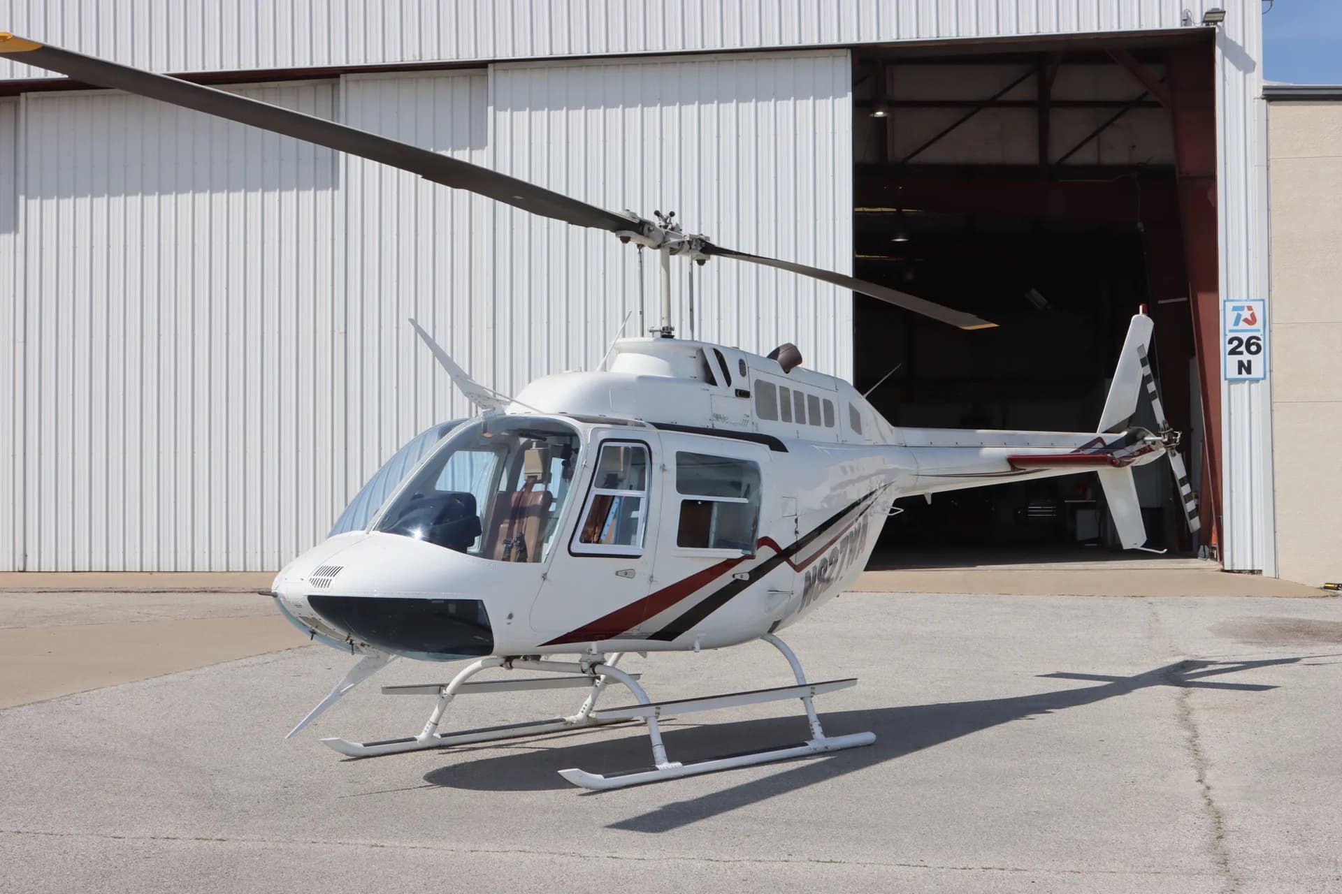 A white Bell 206 JetRanger helicopter with red and black stripes is parked on asphalt in front of an open hangar door.