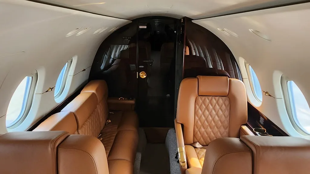 A view down the aisle of a business jet cabin, showing brown leather seats and a dark partition at the rear.