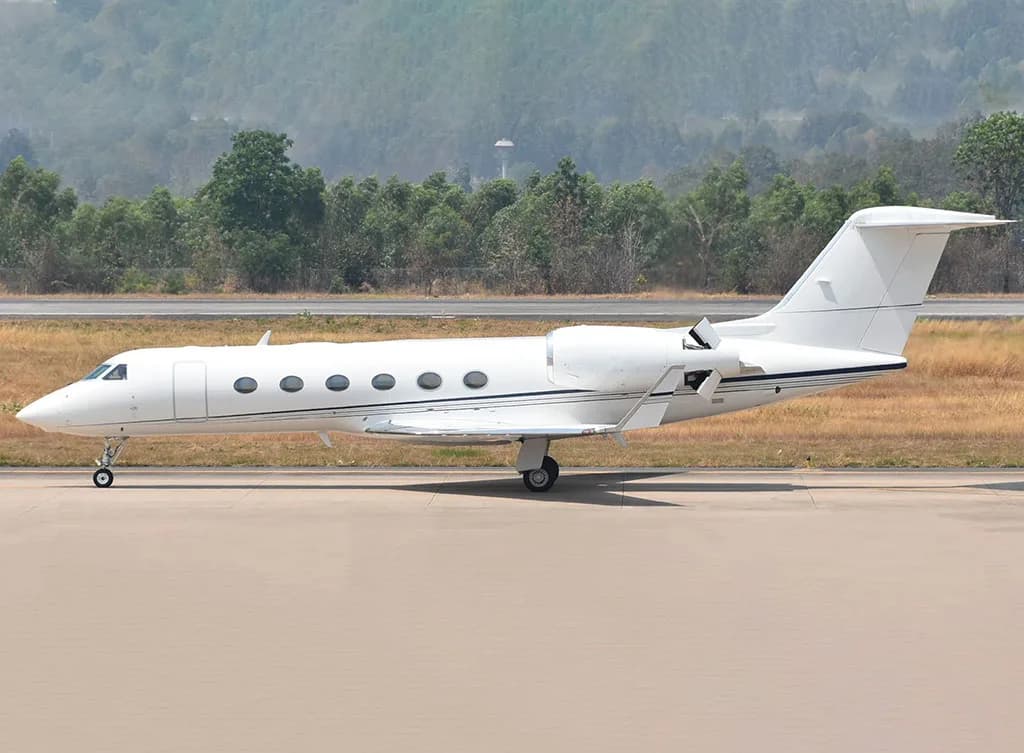 A white Gulfstream business jet parked on an airport tarmac with trees and hills in the background.