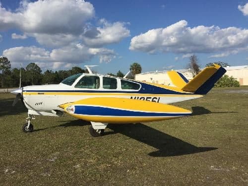 A Beechcraft Bonanza aircraft with a white, yellow, and blue paint scheme is parked on the grass.