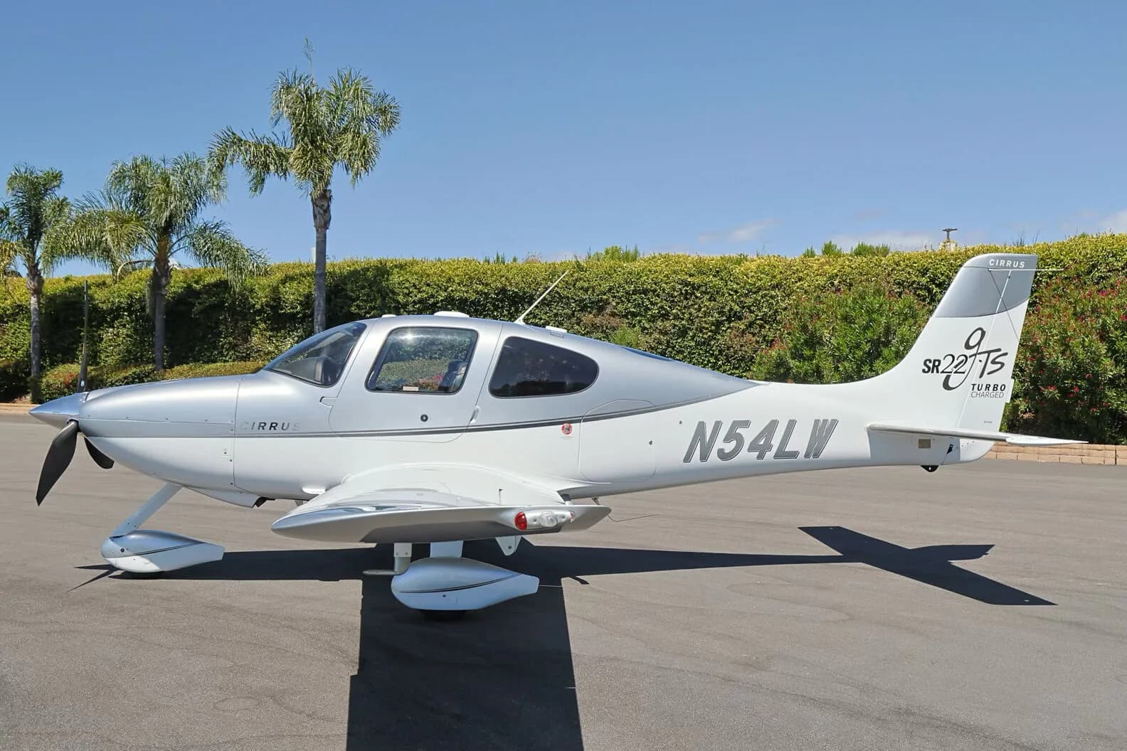 Side view of a silver and white Cirrus SR22T (N54LW) parked on an tarmac with palm trees in the background.